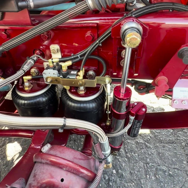 Close-up of a red agricultural tractor or heavy equipment chassis with black air suspension bags, metal hoses, and mechanical components on an outdoor paved surface.