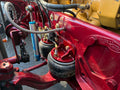 Close-up of a red agricultural tractor or heavy equipment chassis with air suspension bags, metal hoses, and a blue shock absorber. A yellow engine block is visible in the upper right, and the equipment is photographed outdoors on pavement.