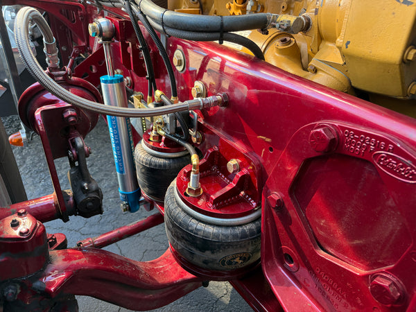 Close-up of a red agricultural tractor or heavy equipment chassis with air suspension bags, metal hoses, and a blue shock absorber. A yellow engine block is visible in the upper right, and the equipment is photographed outdoors on pavement.