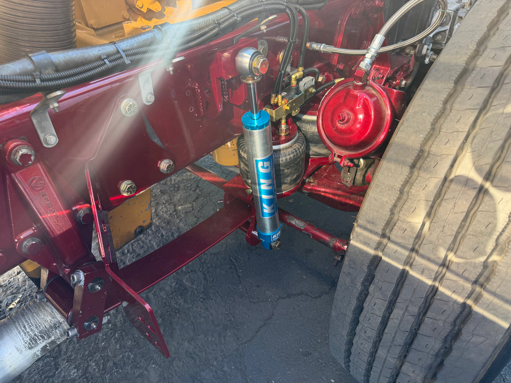 A close-up of a truck's rear frame area showing a blue KYB shock absorber mounted vertically, with red painted metal components and a large tire visible on the right.