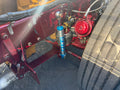 A close-up of a truck's rear frame area showing a blue KYB shock absorber mounted vertically, with red painted metal components and a large tire visible on the right.