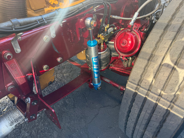 A close-up of a truck's rear frame area showing a blue KYB shock absorber mounted vertically, with red painted metal components and a large tire visible on the right.