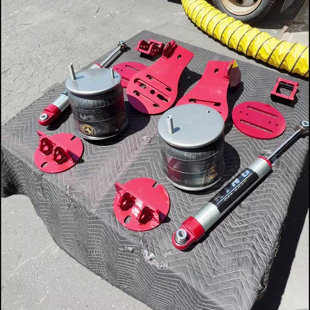 A set of used automotive suspension parts, including two silver coilover bumpers, a shock absorber, and red plastic shims and brackets, displayed on a black textured tablecloth with a red truck in the background.