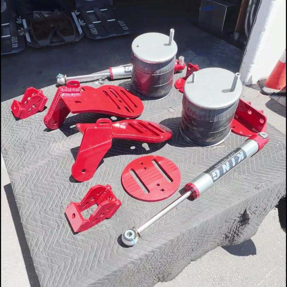 A set of red and silver suspension and shock absorber parts arranged on a gray textured mat outdoors on pavement.