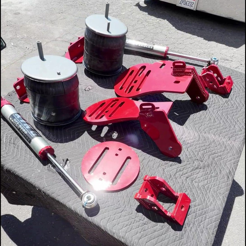 A set of heavy-duty truck suspension parts, including red metal brackets, black and red shock absorbers, and a silver coil spring, laid out on a gray protective mat on asphalt.