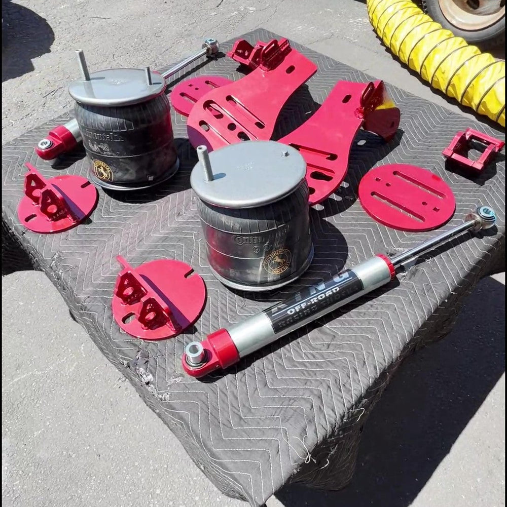 A set of red and silver suspension and shock absorber parts arranged on a black textured mat on the ground, with a red vehicle and a yellow coiled hose visible in the background.