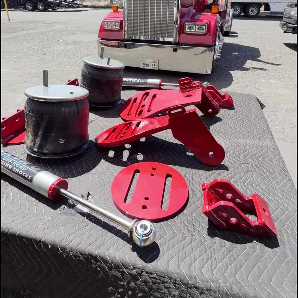 A set of red and silver heavy-duty suspension and shock absorber parts arranged on a black quilted mat in a sunny outdoor lot, with a red semi-truck parked in the background.