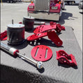 A set of red and silver heavy-duty suspension and shock absorber parts arranged on a black quilted mat in a sunny outdoor lot, with a red semi-truck parked in the background.