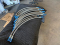 A set of curved stainless steel hydraulic hoses with blue anodized metal fittings, arranged on a black quilted moving blanket in a workshop environment.