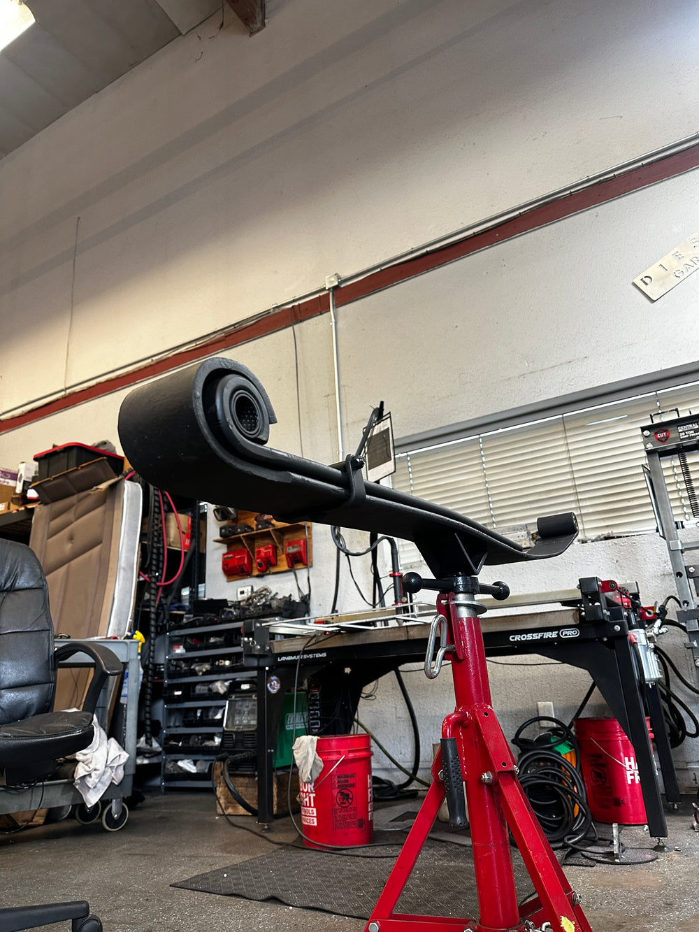 Heavy duty truck leaf spring on jack stand in repair shop with tools and equipment in background.
