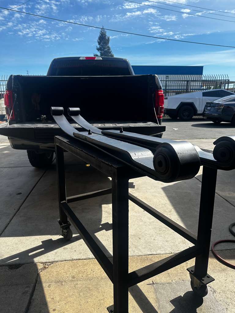 Black metal stand with wheels on a concrete surface, with a truck in the background. leaf springs